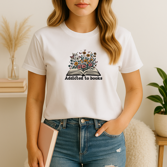A young woman stands in a softly lit reading nook wearing a white bookish t shirt printed with a colorful illustration of an open book bursting into wildflowers and fluttering butterflies above the caption “Addicted to books”; she pairs it with light-wash jeans and holds a novel, with pampas grass, stacked books, and a potted plant styling the neutral background.