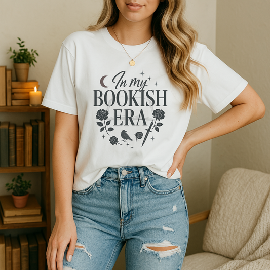 A young woman stands in a cozy reading nook wearing a white bookish shirt printed with “In My Bookish Era” in stylized black lettering, framed by rose blooms, a perched crow, a dagger, crescent moon, and sparkles; behind her, a wooden bookshelf of vintage books, a lit candle, potted greenery, and a cream armchair complete the warm, inviting scene.