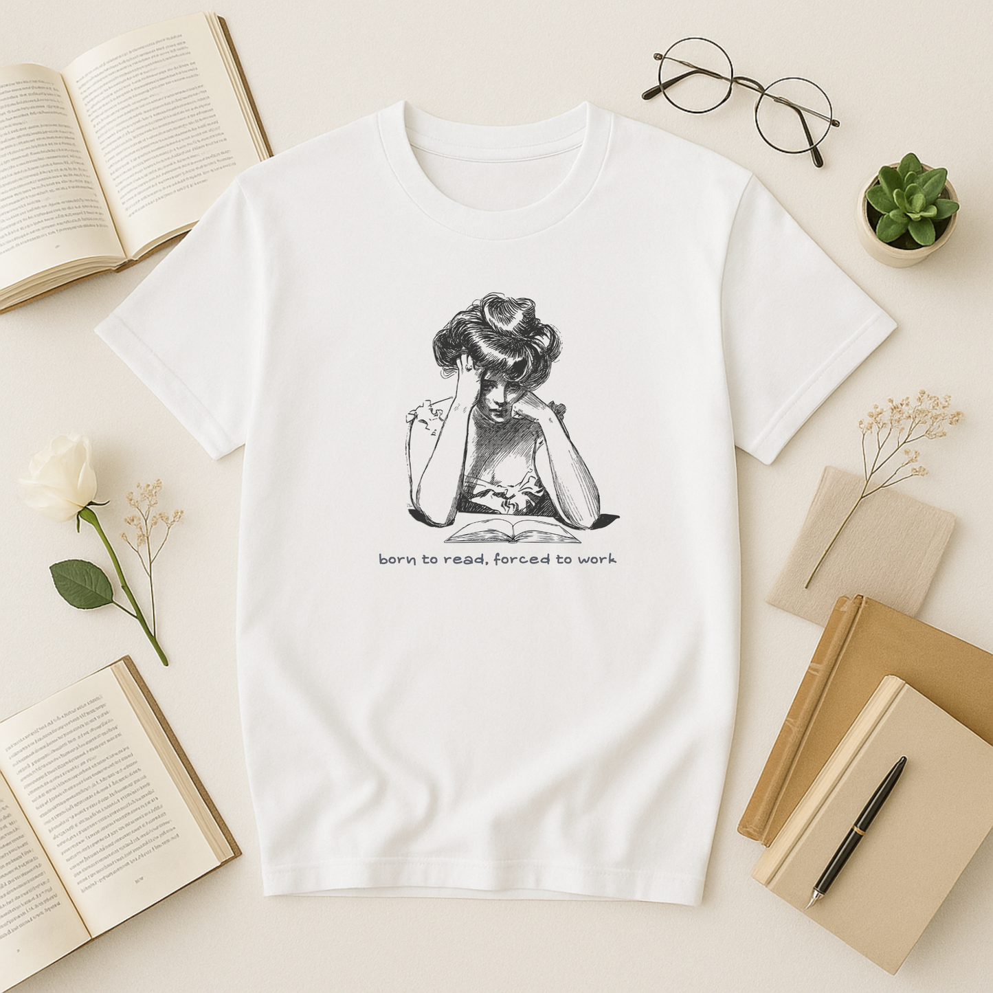 A crisp white bookish t shirt laid flat on a pale surface, printed with a vintage black-and-white illustration of a reader slumped over an open book under the caption “born to read, forced to work”; styled alongside an open novel, round gold-rimmed glasses, a small succulent, a single white rose with baby’s-breath sprig, a linen napkin, a stack of beige notebooks, and a fountain pen.
