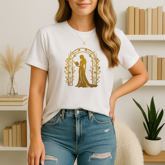 A young woman stands in a softly lit reading nook wearing a white bookish t shirt printed in gold with an elegant silhouette of a woman reading beneath an arch of flowering vines; she pairs it with light-wash jeans and simple jewelry, framed by neutral-toned bookshelves and pampas grass.
