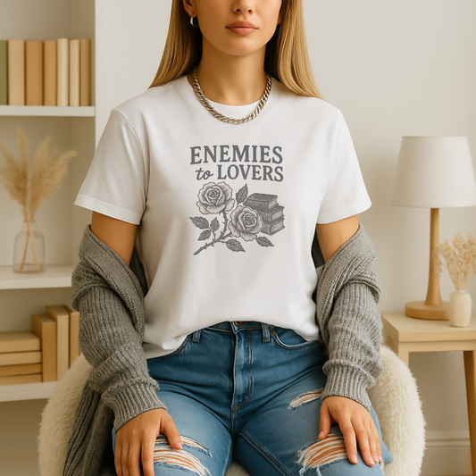 A young woman seated in a softly lit reading nook wears a white book lover t shirt printed with the words “Enemies to Lovers” above a detailed black-and-white illustration of two blooming roses intertwined with a small stack of books; she’s layered in a cozy gray cardigan and distressed jeans, framed by a cream lamp, dried pampas grass, and a shelf of neutral-toned books in the background.








