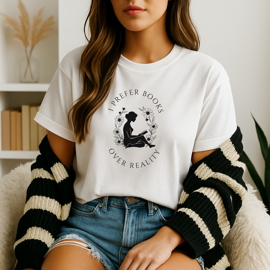 A young woman sits in a cozy reading nook wearing a white book lover t shirt printed with a black silhouette of a seated reader framed by a floral wreath and the words “I Prefer Books Over Reality,” her striped cardigan draped over her arms against a backdrop of neutral-toned bookshelves and pampas grass.