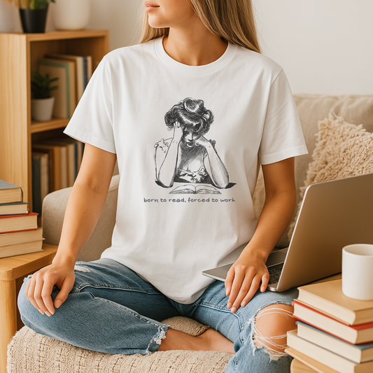 A young woman sits cross-legged on a cozy armchair wearing a white bookish t shirt printed with a vintage black-and-white illustration of a reader propping their head over an open book, captioned “born to read, forced to work,” with a laptop and stacks of books framing the neutral-tone reading nook.







