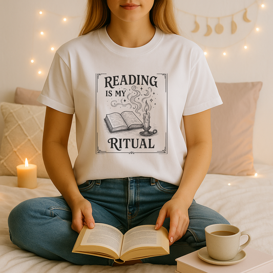 A young woman sits cross-legged on a bed in a softly lit, cozy nook, wearing a white book lover t shirt printed with a vintage-style “Reading Is My Ritual” design of an open book and a smoking candle; she holds an open novel in her lap, surrounded by a lit pillar candle, fairy lights, a moon-phase garland, plush pillows, and a steaming cup of coffee.