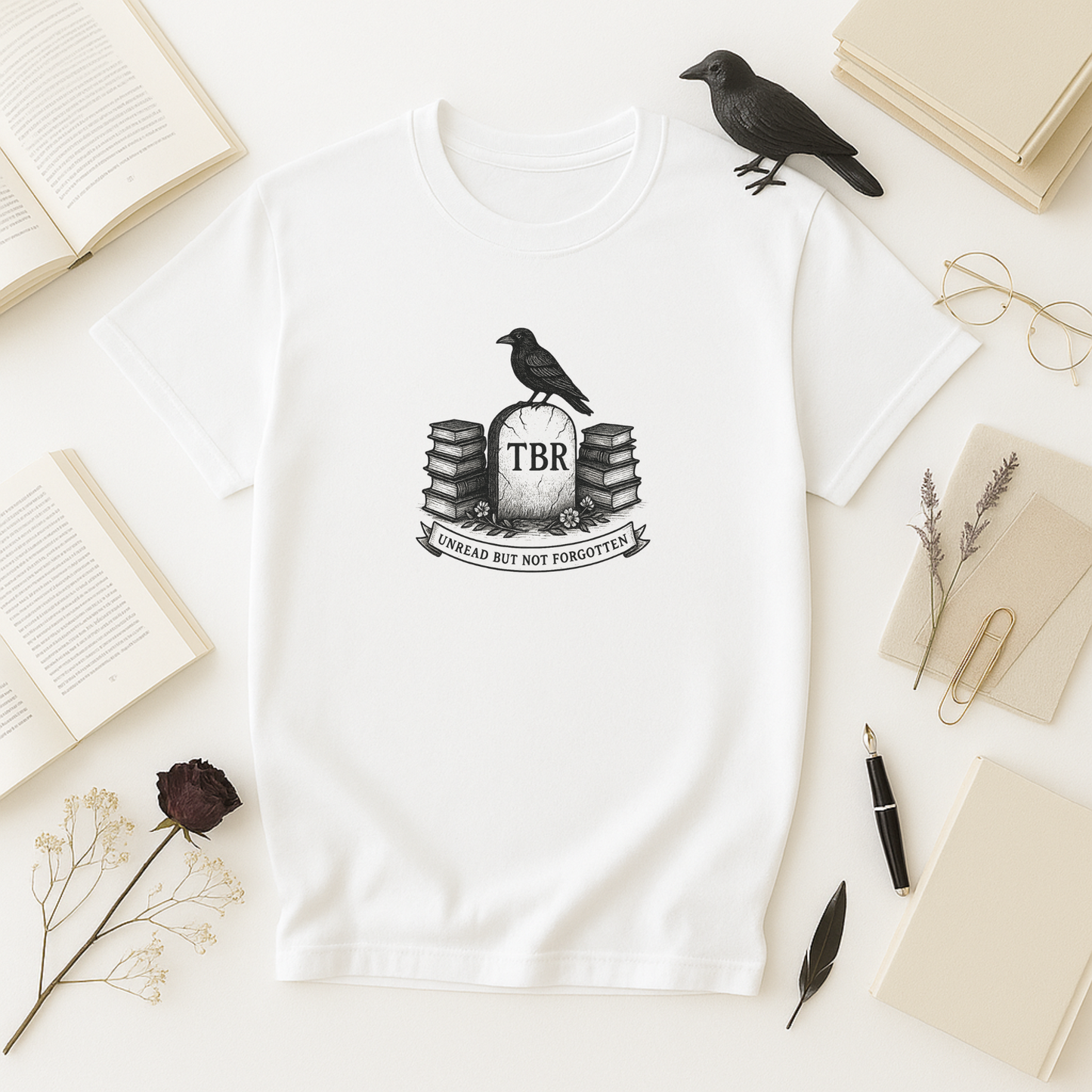 A crisp white bookish t shirt laid flat on a pale surface, featuring a black-and-white “TBR” tombstone design flanked by stacks of books and topped by a perched crow; opened books, a crow figurine, gold-rimmed glasses, scattered dried florals, a fountain pen, and a single dark rose frame the scene.