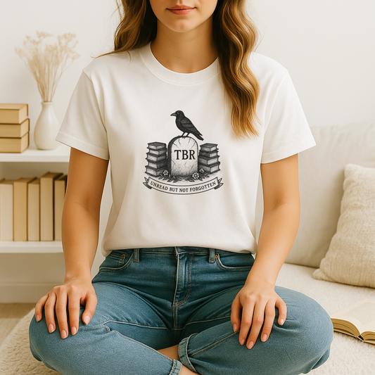 A young woman sits cross-legged on a soft rug in a cozy corner, wearing a white bookish t shirt printed with a black-and-white “TBR” tombstone flanked by stacks of books and topped by a perched crow, with a neutral-toned bookshelf, dried florals, and a cream sofa pillow softly framing the scene.